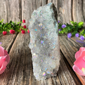 A photograph of a narrow Angel Aura Amethyst Crystal Cluster against a soft, floral backdrop. The cluster features purple amethyst crystals adorned with a delicate iridescent coating, emitting a shimmering rainbow sheen in the light.